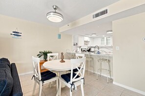A dining area with a round table, white chairs, a ceiling fan, and a kitchen in the background. - Modern Ocean View Escape (Indian Shores)