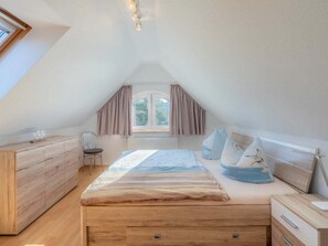 A bedroom with a sloped ceiling, a bed with a striped bedspread, a wooden dresser, a chair, and a window with curtains. - Holiday Apartment With 1 Bedroom (Heringsdorf)
