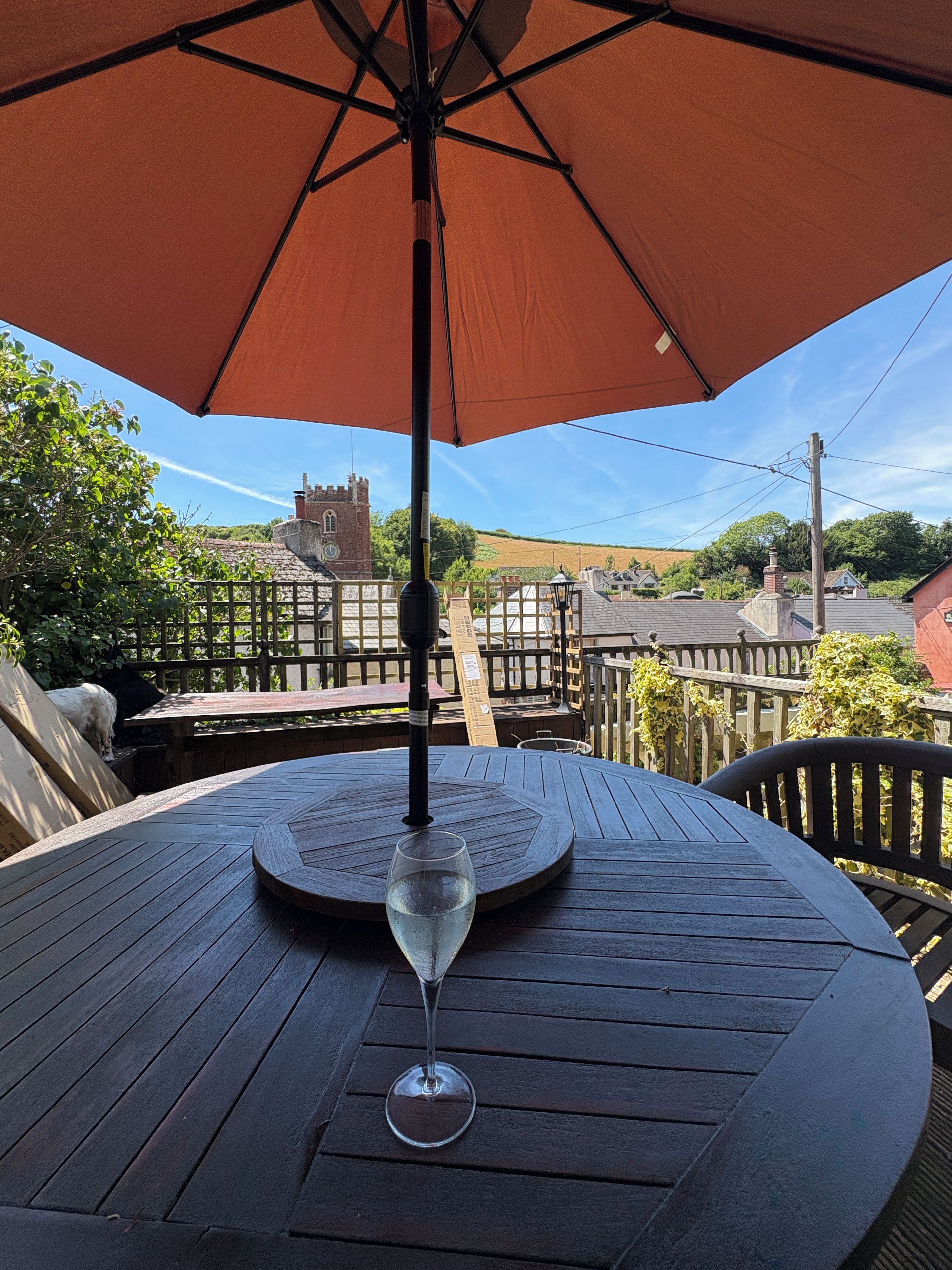 A round wooden table with a glass of water under an umbrella, overlooking a village with traditional buildings and a tower.