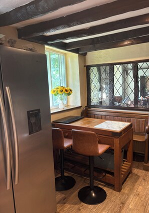 A kitchen with a stainless steel refrigerator, wooden bar stools, a wooden table, and a window with a view of greenery. - Beautiful 400 yr old thatch house.Sleeps 11, dog friendly,close to beaches,moors (Combeinteignhead)