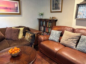 A living room with a brown leather sofa, a wooden coffee table with oranges, a bookshelf, and framed artwork on the walls. - Beautiful 400 yr old thatch house.Sleeps 11, dog friendly,close to beaches,moors (Combeinteignhead)