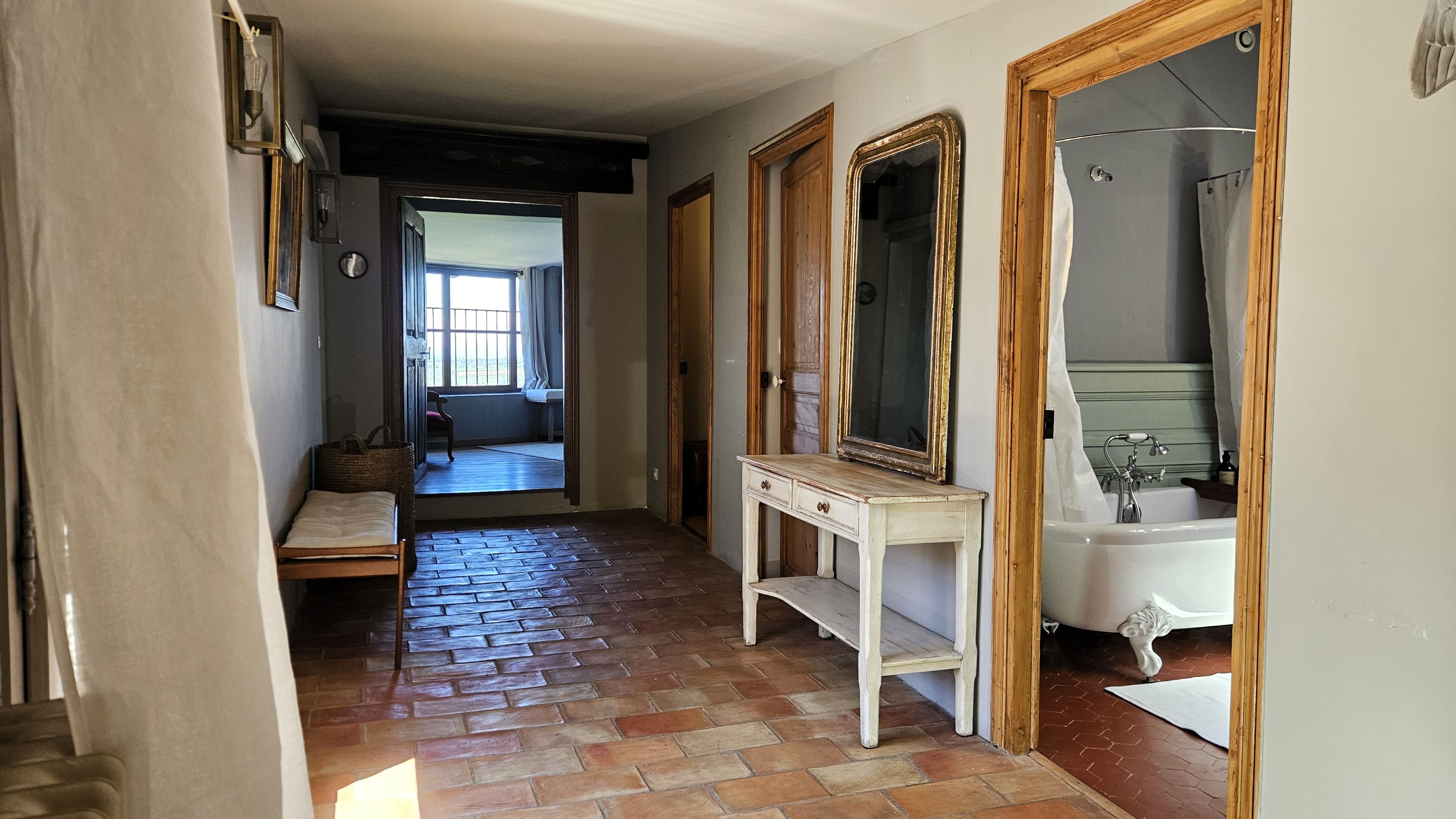 A hallway with a wooden floor, a bench, a table with a mirror, and a bathroom with a bathtub.
