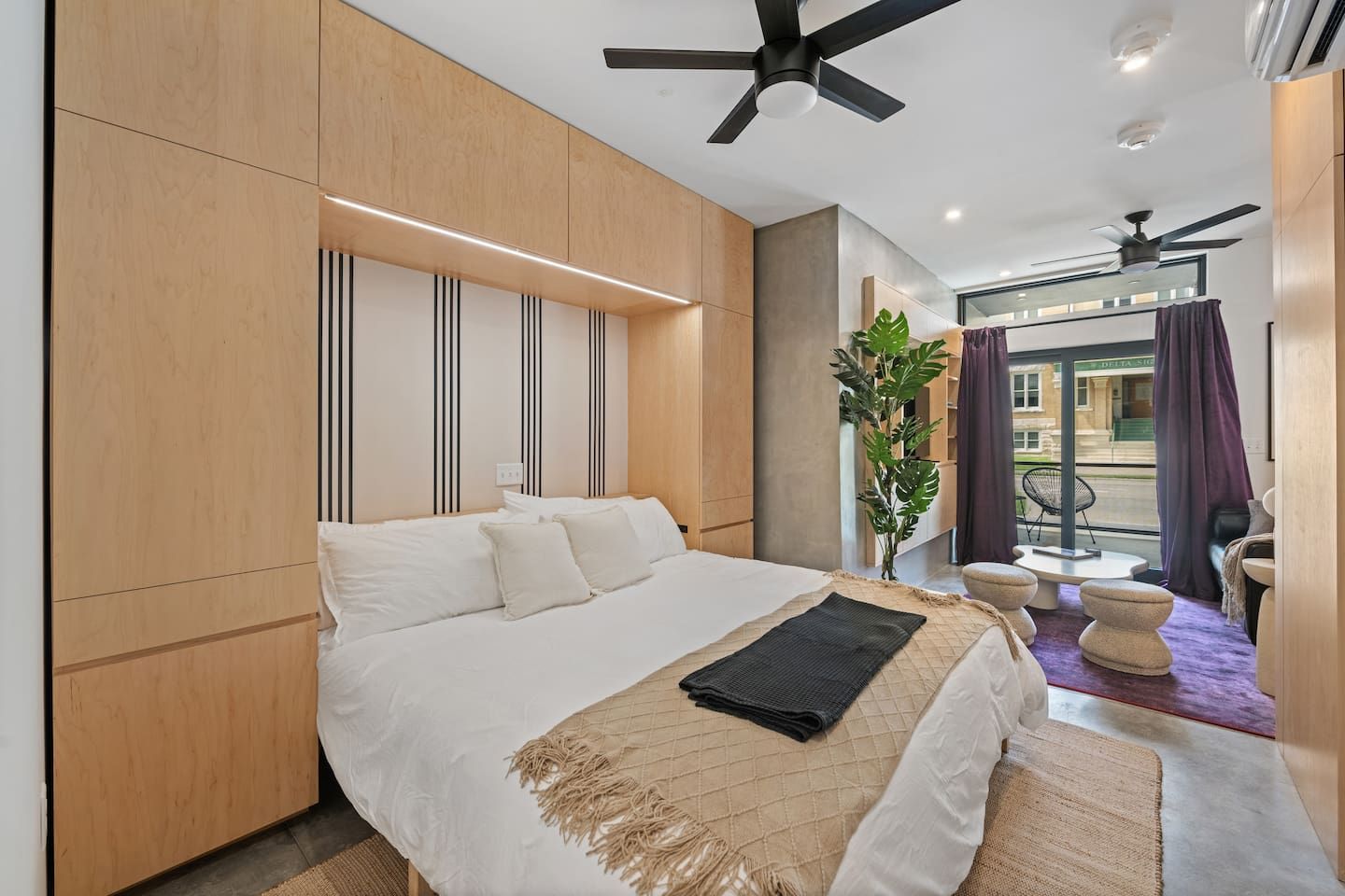 A modern hotel room with a large bed, a ceiling fan, a view of the street, and a balcony with seating.