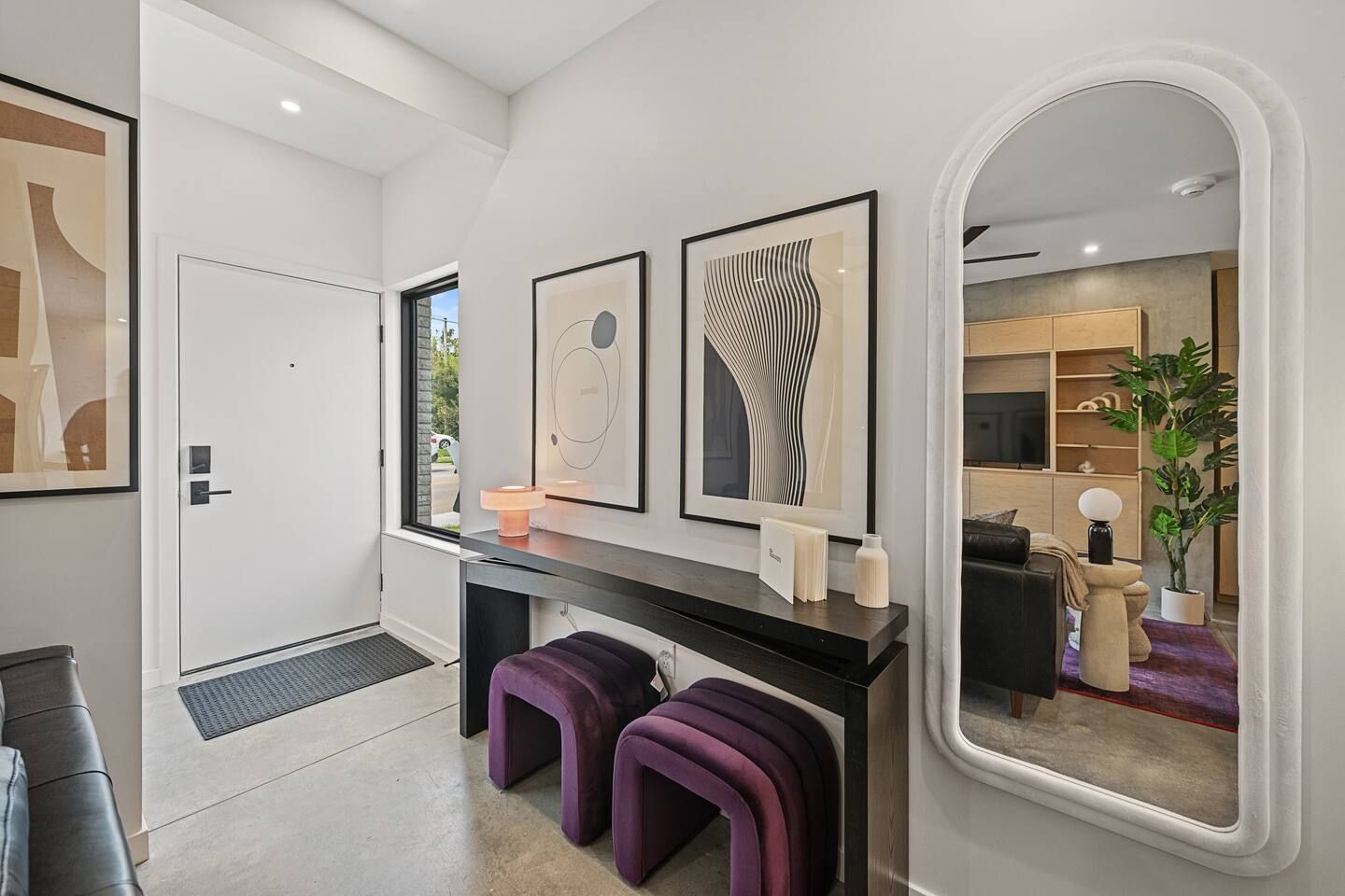 A modern interior with a white door, a console table with artwork, purple stools, and a large mirror reflecting a living area.