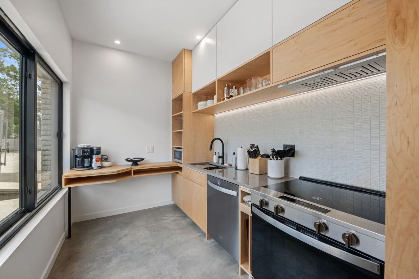 A modern kitchen with a built-in oven, stainless steel appliances, wooden shelves, and a window providing natural light.