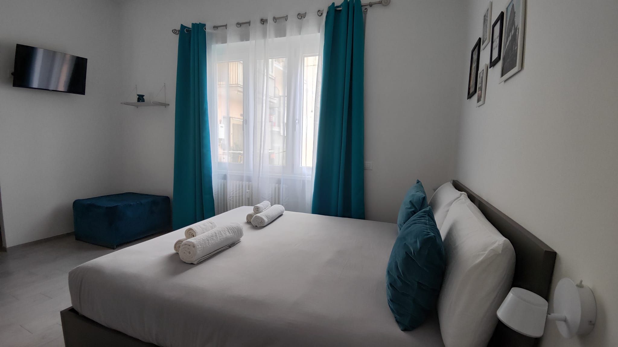 Ein ordentlich bezogenes Bett mit weißen Leinen und türkisfarbenen Kissen, ein blauer Hocker, ein Flachbildfernseher an der Wand und ein Fenster mit türkisfarbenen Vorhängen.