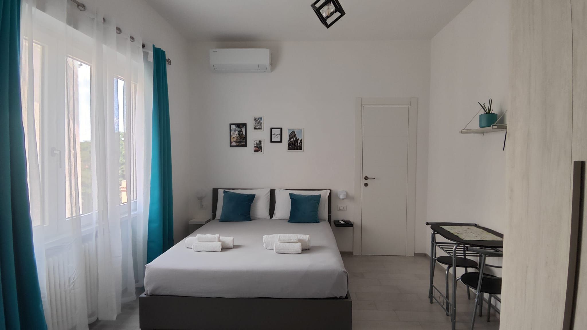 Une chambre d’hôtel avec un lit, deux oreillers, une table de chevet, un bureau, une chaise, une porte et une fenêtre avec des rideaux.