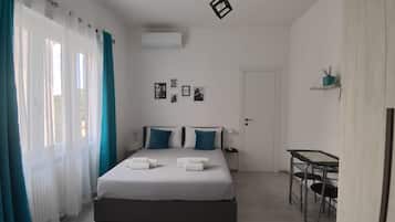 Une chambre d’hôtel avec un lit, deux oreillers, une table de chevet, un bureau, une chaise, une porte et une fenêtre avec des rideaux.