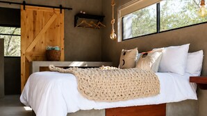 A bedroom with a concrete bed, wooden headboard, a hanging light fixture, and a window with a view of trees. - Luxury Bush Villa | Private Pool, Hot Tub in Marloth at the Kruger National Park (Marloth Park)