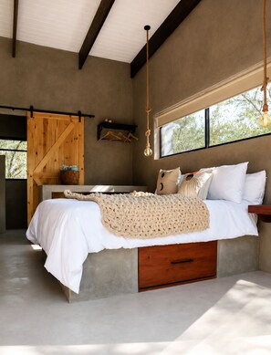 A bedroom with a concrete bed, wooden headboard, a hanging light fixture, and a window with a view of trees. - Luxury Bush Villa | Private Pool, Hot Tub in Marloth at the Kruger National Park (Marloth Park)