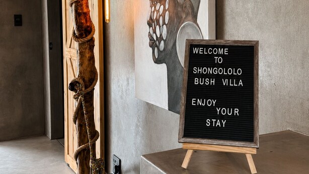 A reception area with a wooden signboard, a wall-mounted light fixture, and a piece of artwork depicting a floral arrangement. - Luxury Bush Villa | Private Pool, Hot Tub in Marloth at the Kruger National Park (Marloth Park)