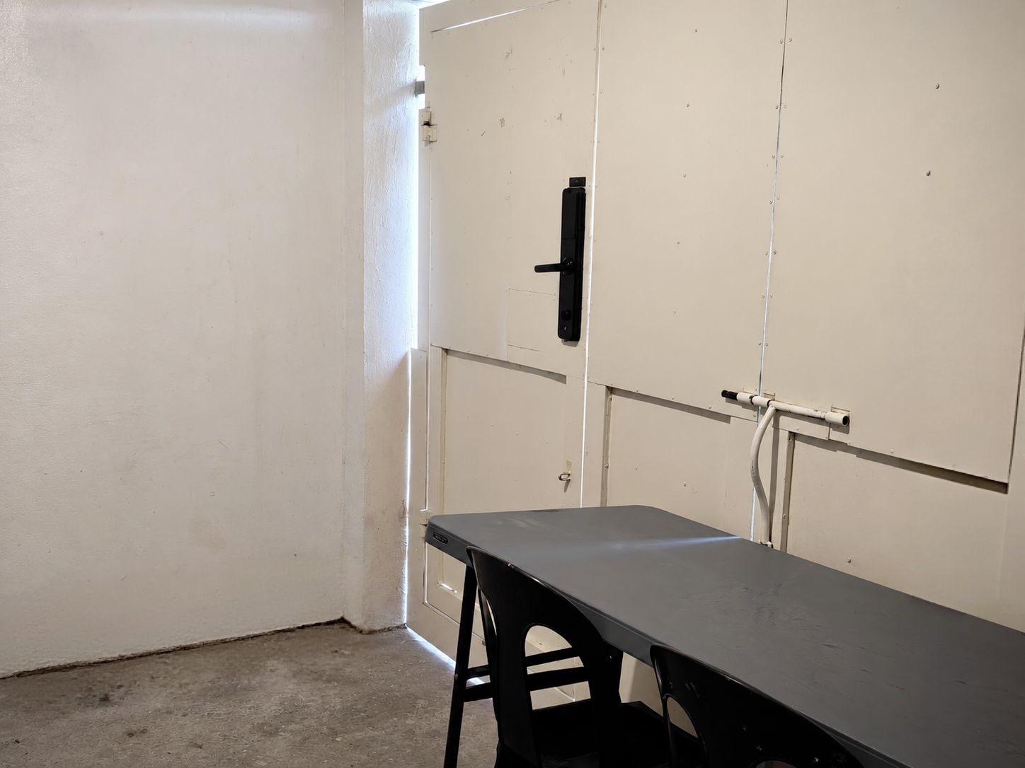 Ein Zimmer mit einem Tisch und Stühlen, eine weiße Tür mit schwarzem Griff und einer sichtbaren Rohrleitung an der Wand.