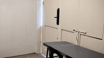 Ein Zimmer mit einem Tisch und Stühlen, eine weiße Tür mit schwarzem Griff und einer sichtbaren Rohrleitung an der Wand.