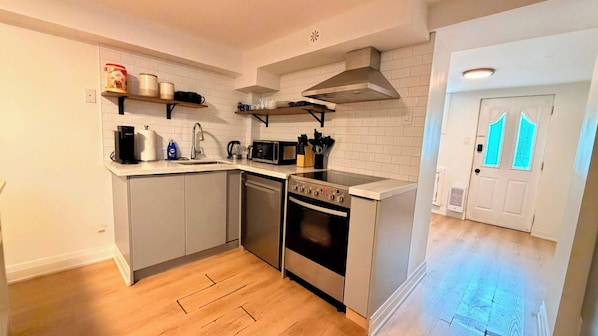 A compact kitchen with a dishwasher, stove, and microwave. There are shelves above the counter holding various kitchen items. - Elegant Trinity Bellwoods 1BR2BA Parkside Retreat (Toronto)