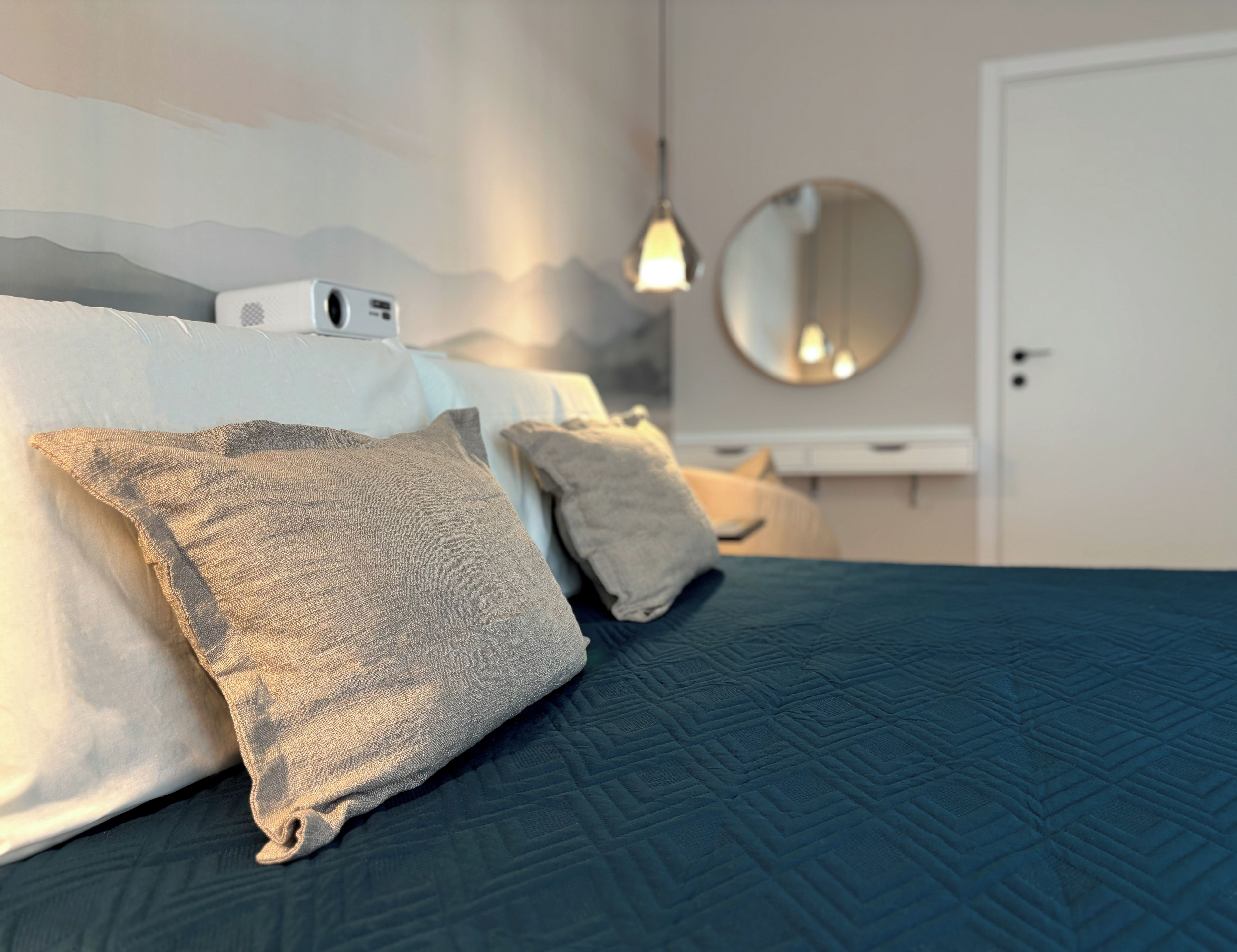 Un lit avec une couverture sombre, deux oreillers, une table de chevet, un miroir fixé au mur et un luminaire suspendu.