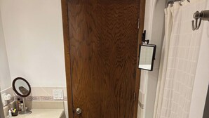 Une salle de bain avec une porte en bois, une baignoire blanche, un rideau de douche, un petit miroir et un tapis beige.