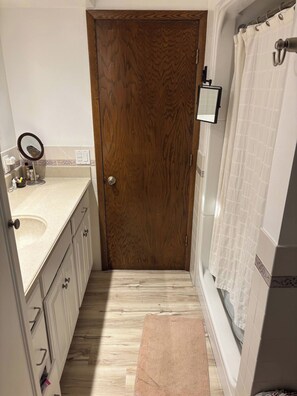 A bathroom with a wooden door, a white bathtub, a shower curtain, a small mirror, and a beige rug. - EAA Gem! Minutes away from the AirVenture grounds. (Oshkosh)