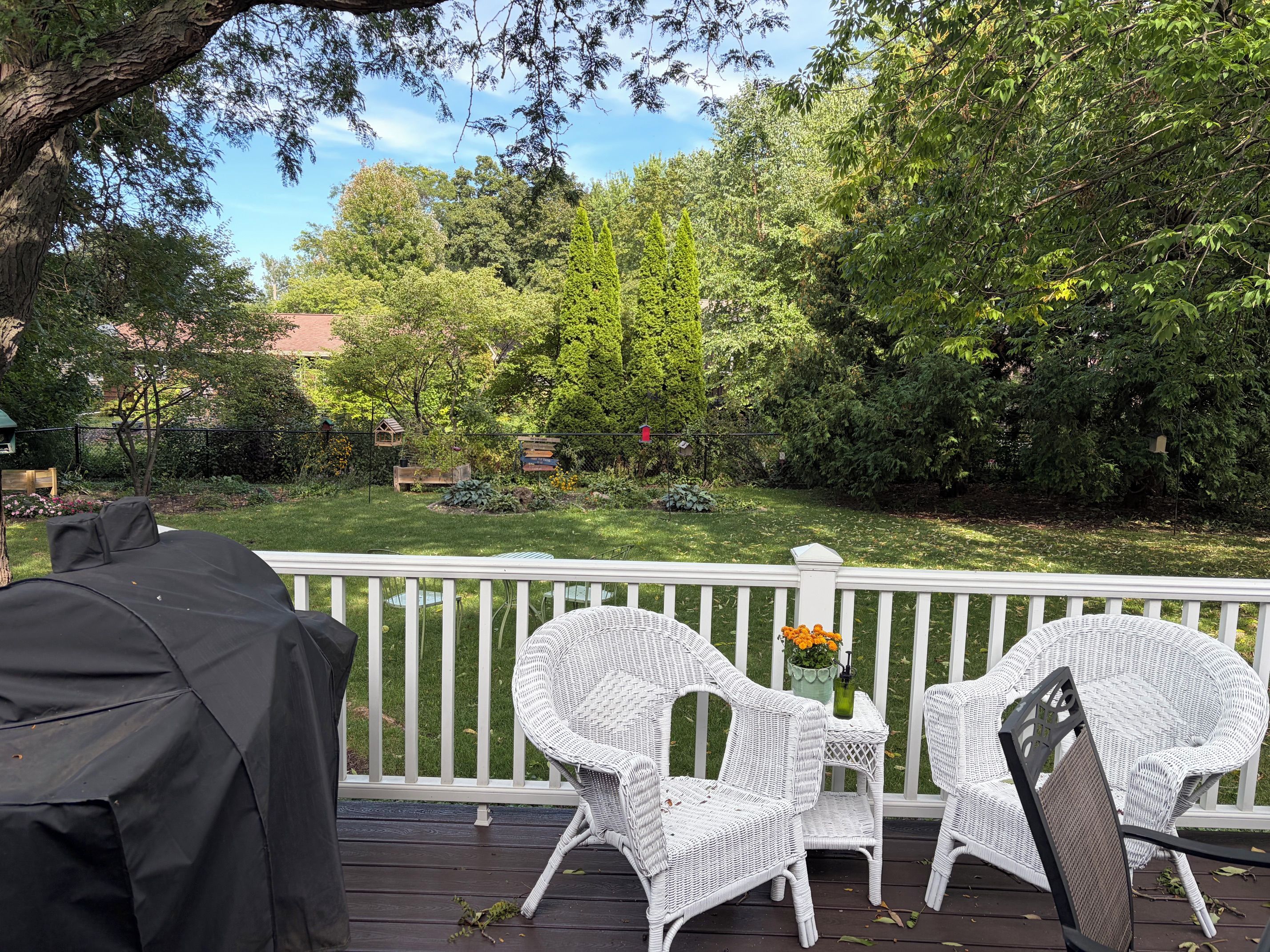 A deck with white wicker chairs and a small table, overlooking a lush garden with a variety of plants and a wooden bench.