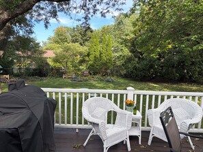 A deck with white wicker chairs and a small table, overlooking a lush garden with a variety of plants and a wooden bench.