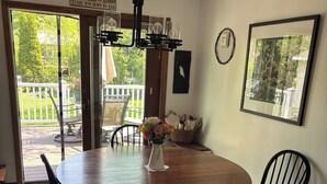 Un coin repas avec une table ronde en bois, quatre chaises noires, un vase avec des fleurs, un luminaire suspendu et une vue sur l’extérieur grâce à une porte vitrée.