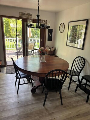 A dining area with a round wooden table, four black chairs, a vase with flowers, a hanging light fixture, and a view of the outdoors through a glass door. - EAA Gem! Minutes away from the AirVenture grounds. (Oshkosh)
