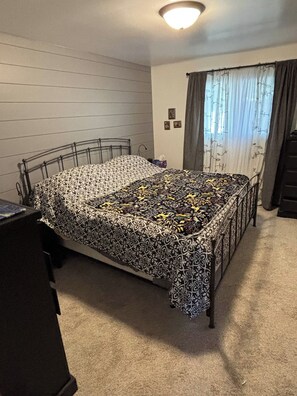 A bedroom with a metal bed frame, a patterned bedspread, a carpeted floor, a dresser, and a window with curtains. - EAA Gem! Minutes away from the AirVenture grounds. (Oshkosh)
