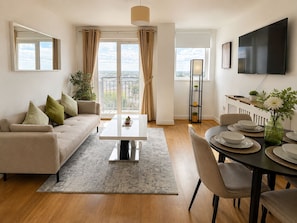 A modern living room with a sofa, coffee table, dining table set for four, and a TV mounted on the wall. - Modern 2 bed 2 bath - 1 Min to Station - Skyline View - Sleeps 5 (Ilford)