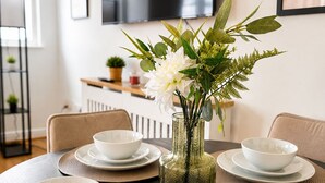 Mesa de jantar posta com tigelas e pratos brancos, um vaso com flores e uma televisão fixada na parede.