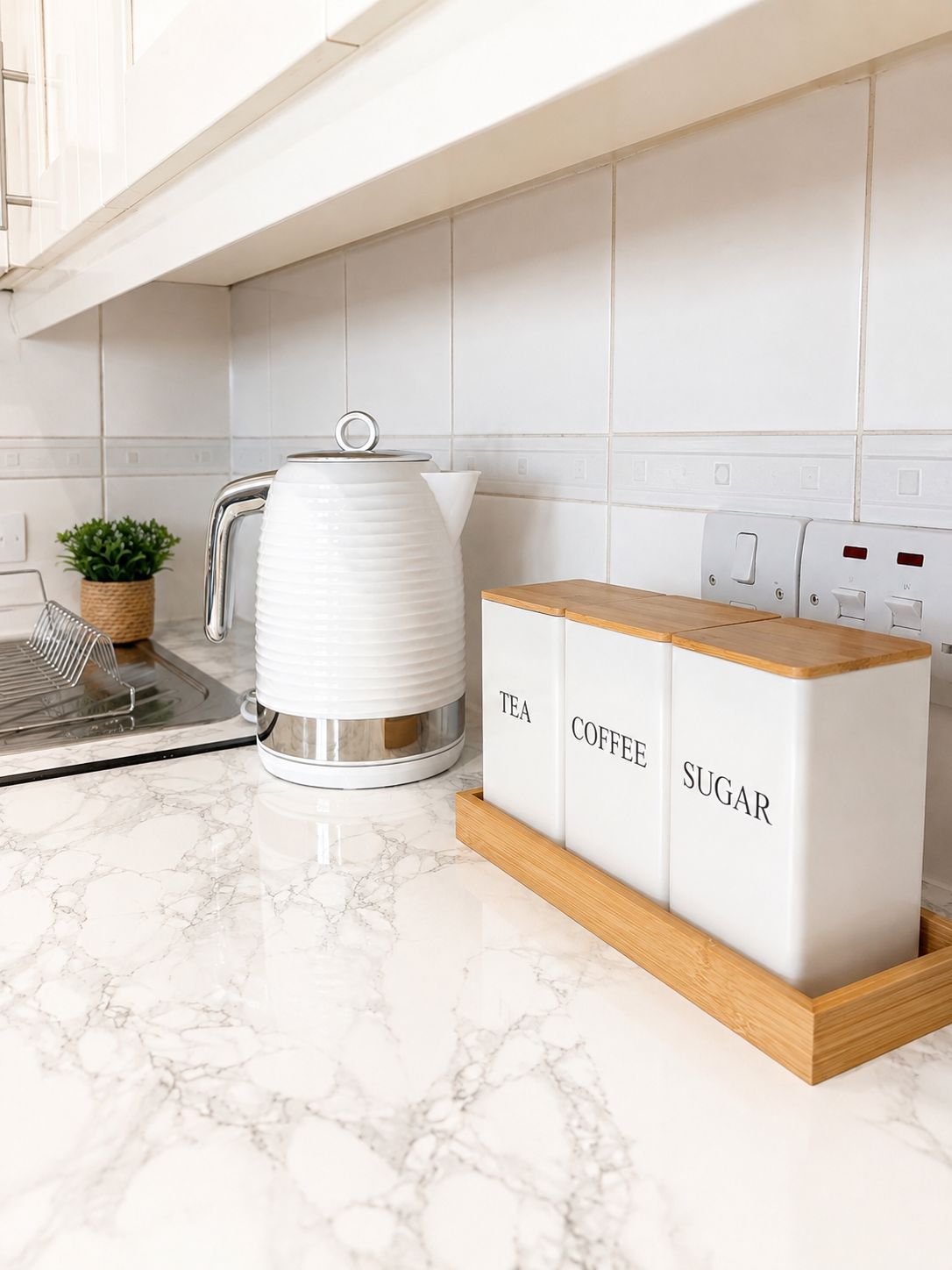 Eine Küche mit einem weißen Wasserkocher, einer Kaffeemaschine und einem Holzschrank mit Fächern für Tee, Kaffee und Zucker.