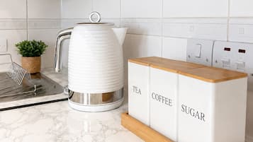 Eine Küche mit einem weißen Wasserkocher, einer Kaffeemaschine und einem Holzschrank mit Fächern für Tee, Kaffee und Zucker.