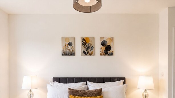 Cama bem arrumada com cabeceira escura, roupa de cama branca e manta de cor mostarda. Duas mesas de cabeceira com luminárias, e três obras de arte penduradas na parede acima da cama.