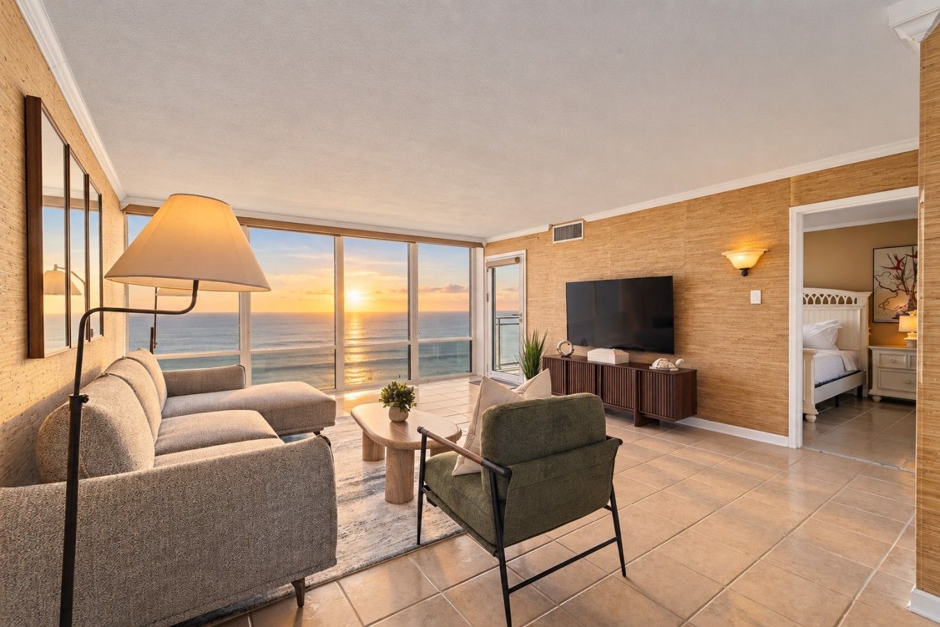 A modern living room with a sofa, armchair, and a TV. There is a large window with a view of the ocean.