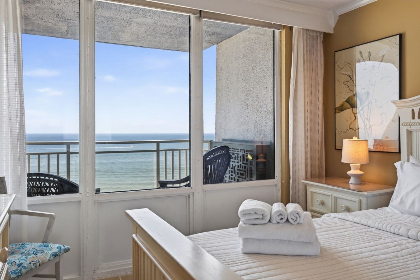 A hotel room with a bed, a chair, a nightstand, a lamp, and a view of the ocean.