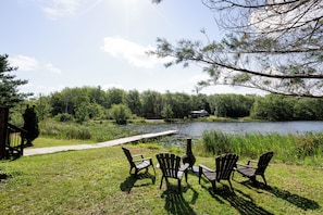 A lakeside outdoor seating area with chairs and a table, surrounded by greenery and a dock extending into the water.