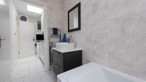 A modern bathroom with a white bathtub, a sink, and a mirror. There is a door leading to another room and a clock on the wall. - Blue Becontree in Goresbrook Village, London (Dagenham)