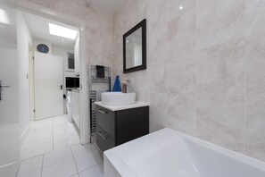 Une salle de bain moderne avec une baignoire blanche, un lavabo et un miroir. Une porte donne accès à une autre pièce et une horloge est accrochée au mur.