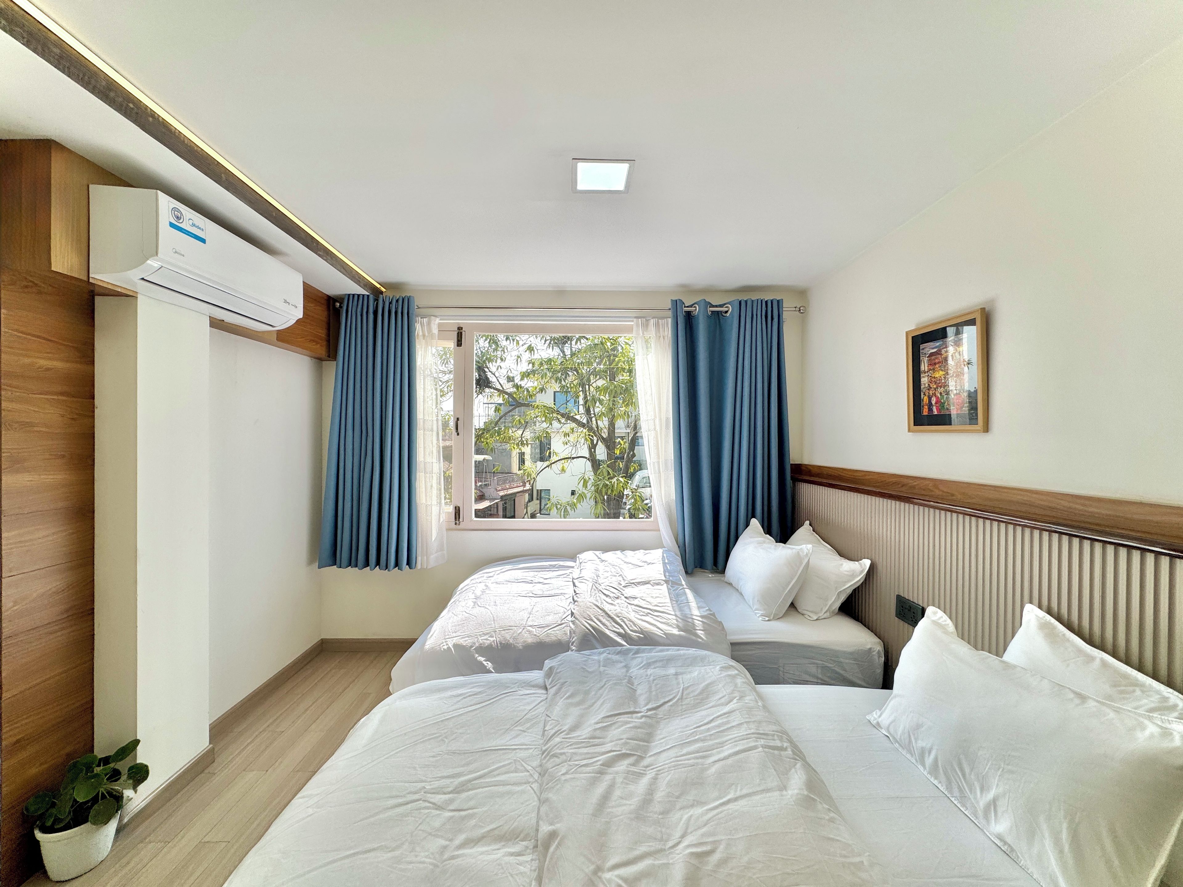 Habitación de hotel con dos camas, una ventana con cortinas, un equipo de aire acondicionado, una planta en maceta y un cuadro en la pared.