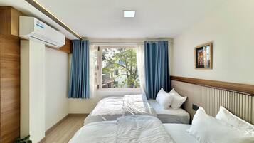 Habitación de hotel con dos camas, una ventana con cortinas, un equipo de aire acondicionado, una planta en maceta y un cuadro en la pared.