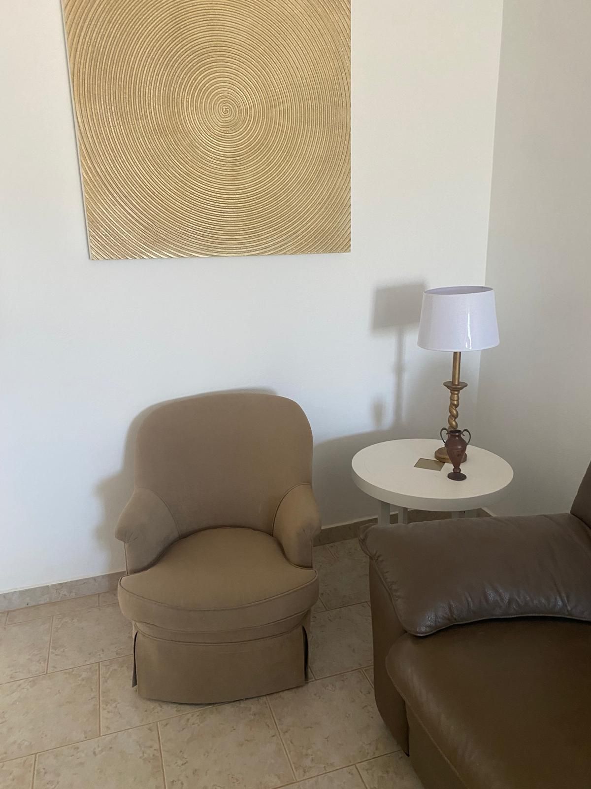 A room with a beige armchair, a small round table with a lamp, and a brown sofa.