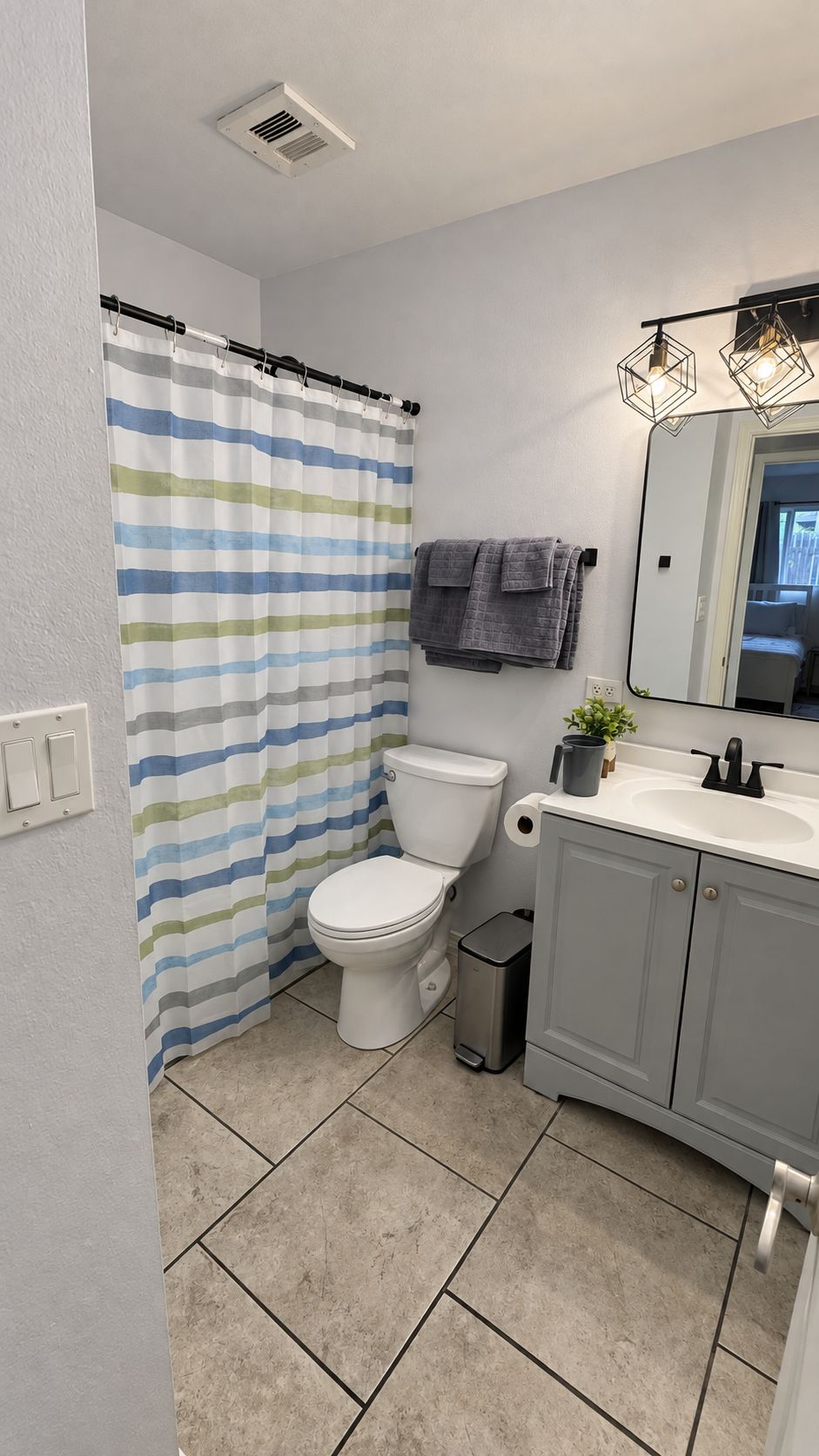 A bathroom with a striped shower curtain, a toilet, a sink with a mirror, and a trash bin.