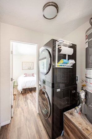 A compact laundry room with a washing machine and dryer, a small sink, and a door leading to a bedroom with a bed and a plant. - Casa Serena (Albuquerque)
