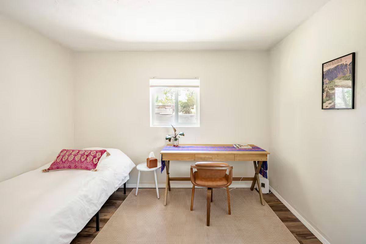 A bedroom with a bed, a desk, a chair, a window, and a wall-mounted picture.