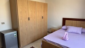 A bedroom with a bed, a wooden wardrobe, a small bedside table, and a compact refrigerator. -  3-bedroom luxury apartment with AC infront of Giza pyramids (Giza Governorate)