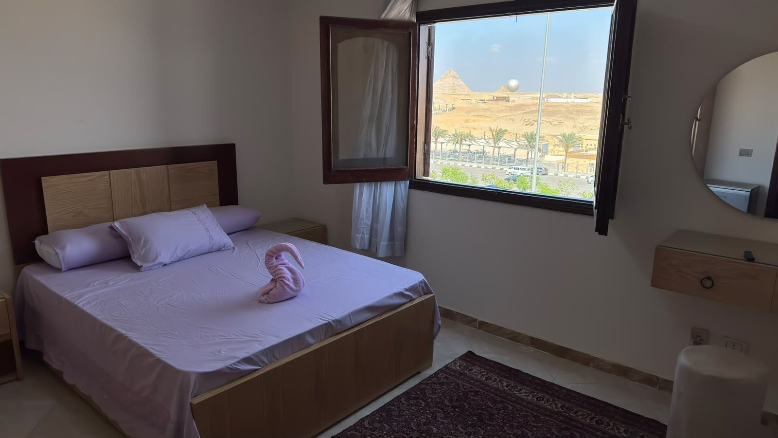 A bedroom with a bed, a window with a view of pyramids, a bedside table, and a mirror.