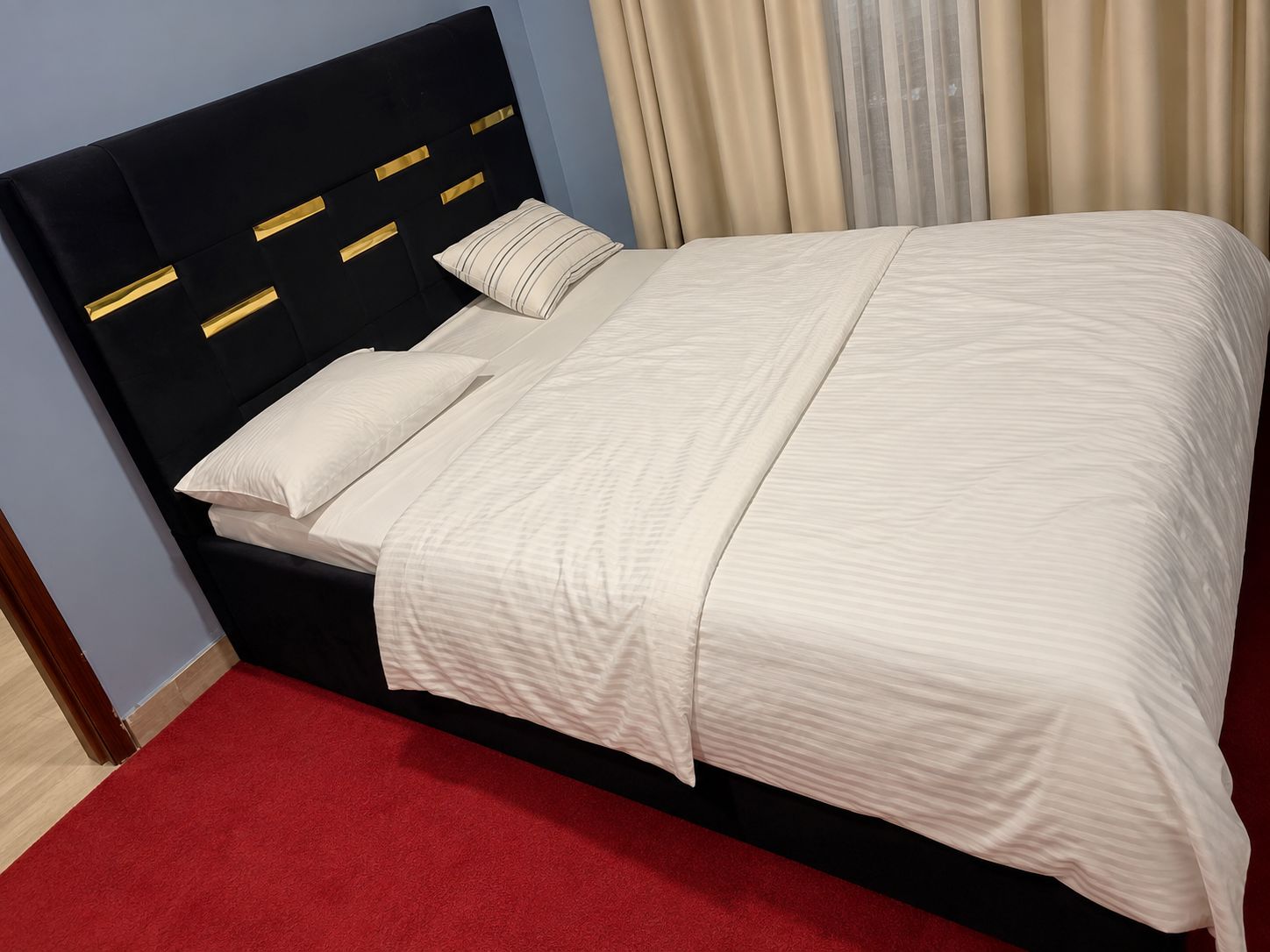 Un lit avec une tête de lit noire et du linge de lit blanc, une moquette rouge et des rideaux beige.