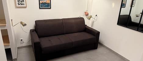 A room with a brown sofa, a mirror, and several framed pictures on the wall.