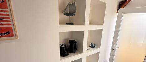 A hotel room with a white wall, a framed poster, a built-in shelf with a lamp, a coffee maker, and a star-shaped container.