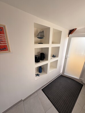 A hotel room with a white wall, a framed poster, a built-in shelf with a lamp, a coffee maker, and a star-shaped container. - Les Gîtes du Corsaire - Le Moussaillon Studio (Broons)