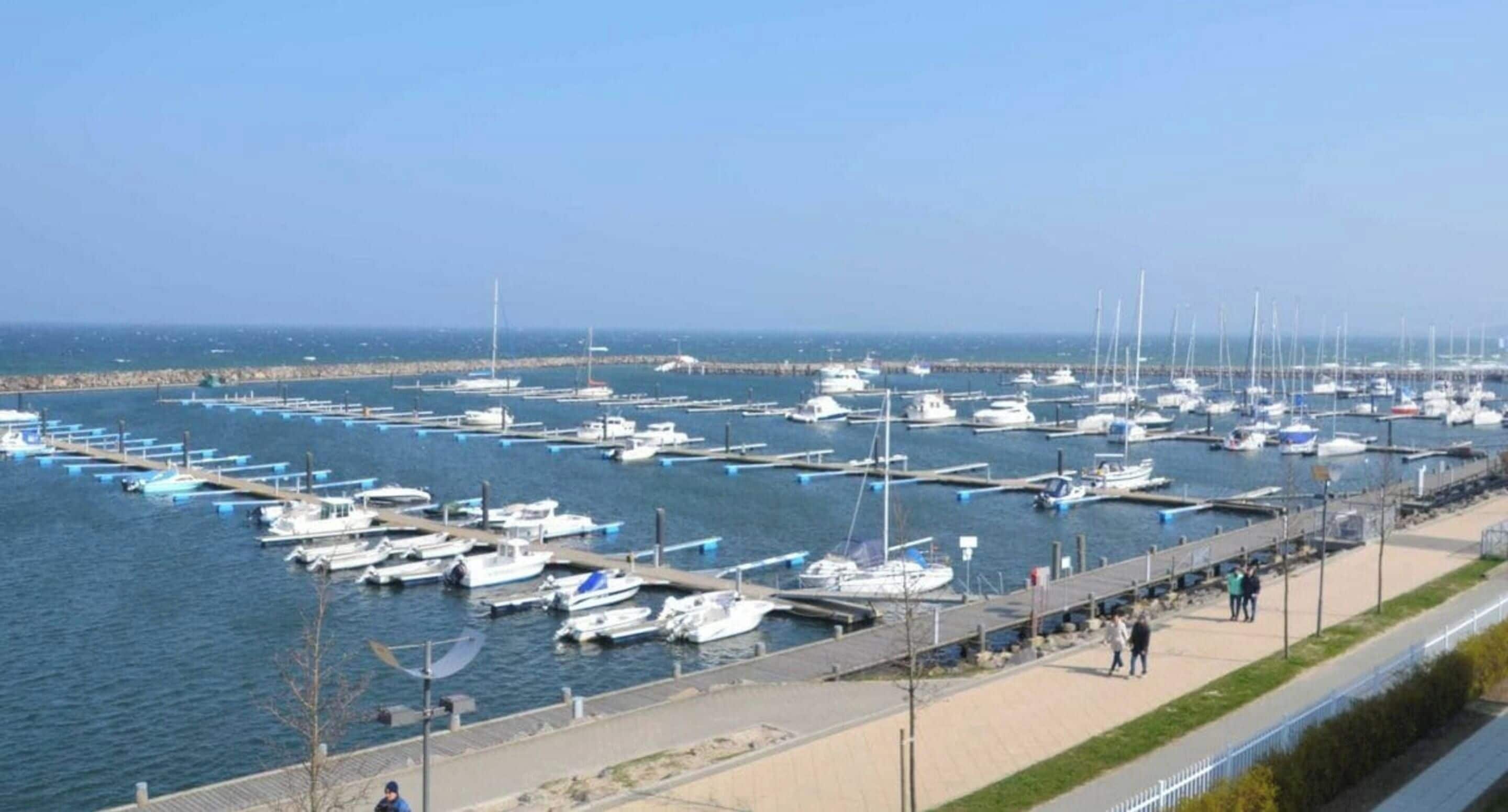 A marina with numerous boats docked at piers, a walkway along the water, and a clear sky.