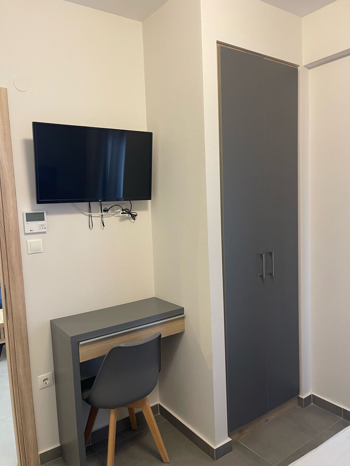 A room with a flat-screen TV mounted on the wall, a desk with a chair, and a wardrobe with two doors.